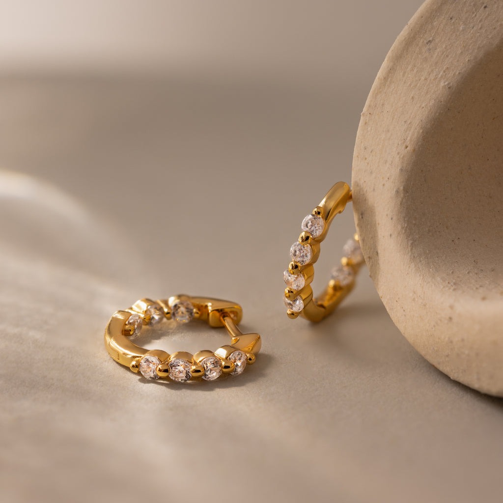 Mini Diamond Beaded Huggies with sparkling diamonds are displayed on a soft beige surface beside a curved stone, highlighting the chic elegance of these modern huggie earrings.