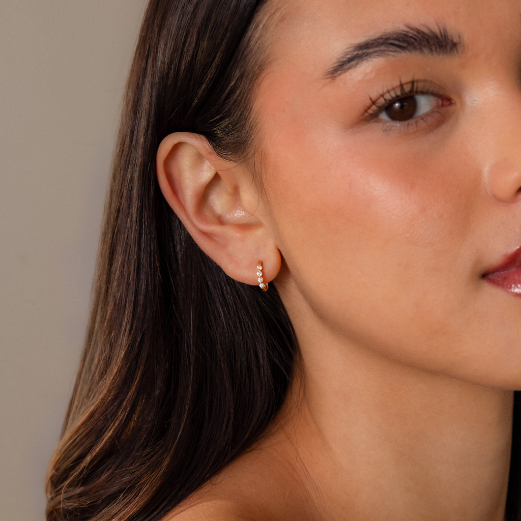 A woman with smooth skin wears the Mini Diamond Beaded Huggies, a modern small gold hoop adorned with sparkling beads, on her left ear.