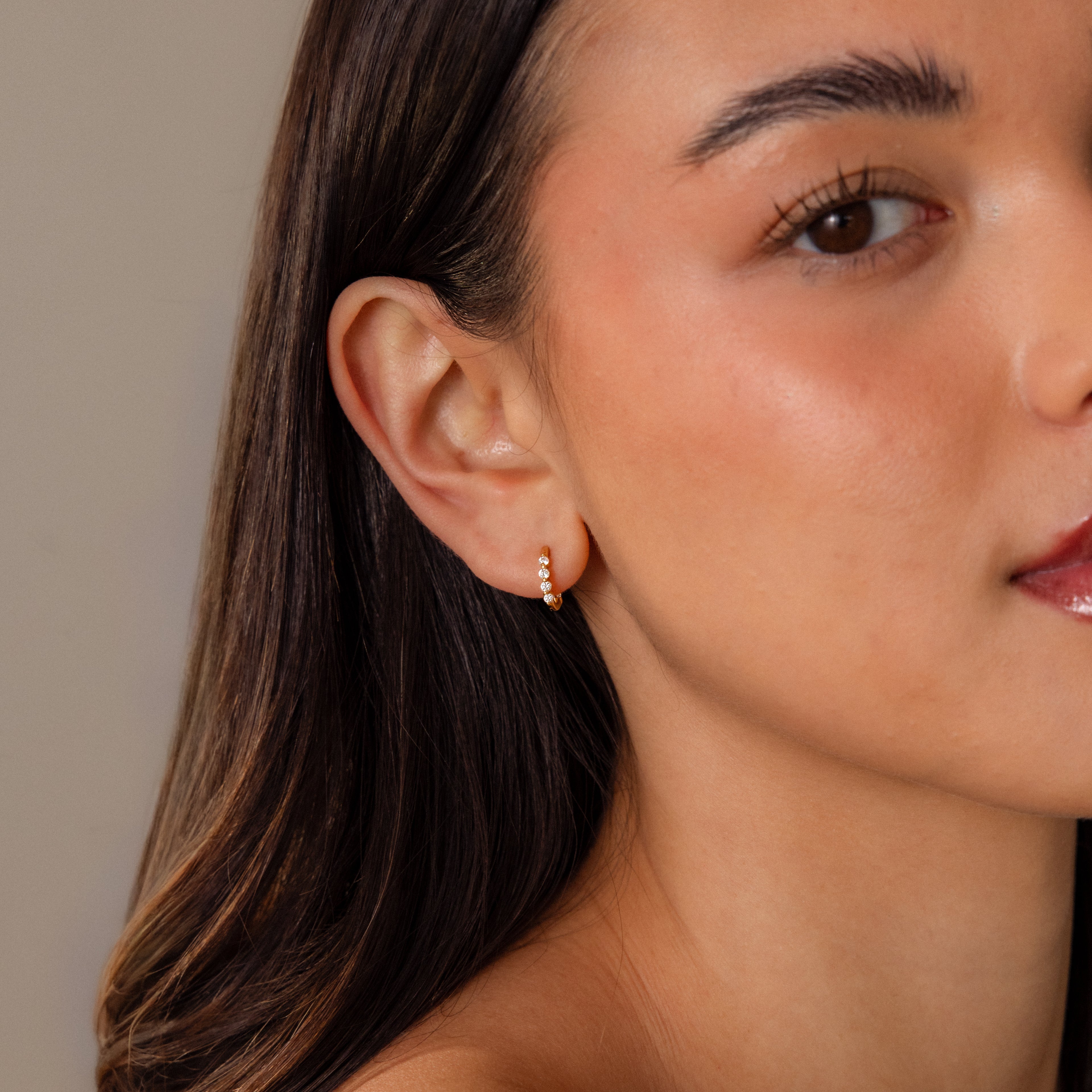 A woman with smooth skin wears the Mini Diamond Beaded Huggies, a modern small gold hoop adorned with sparkling beads, on her left ear.