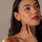 A woman with long dark hair touches her neck, wearing Mini Birthstone Beaded Huggies and natural makeup.