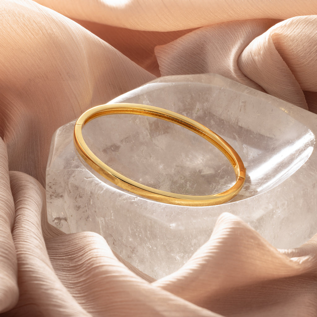 The Classic Bangle in 18K Gold rests on a clear crystal and soft peach fabric—a chic, minimalist accessory perfect for those who love elegant jewelry.