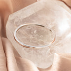 The Classic Bangle in Sterling Silver adds a chic touch, displayed on a large clear crystal and nestled among soft beige fabric folds.