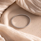 The Classic Bangle is displayed on a textured beige surface with sheer cream-colored fabric draped around it, adding an effortless touch of chic style.