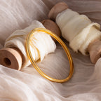 The Classic Bangle, a chic jewelry piece, is elegantly displayed on soft cream fabric and wooden spools.