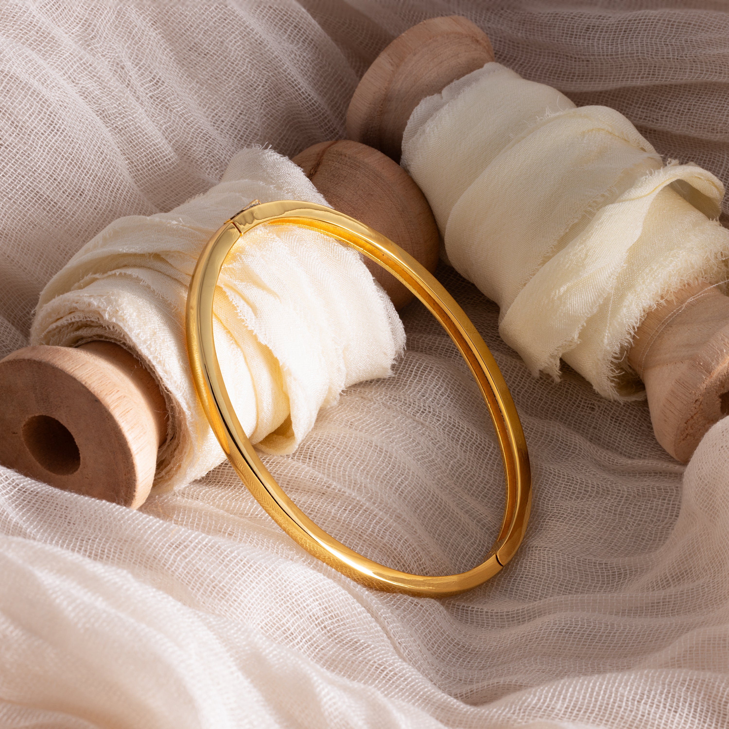 The Classic Bangle, a chic jewelry piece, is elegantly displayed on soft cream fabric and wooden spools.