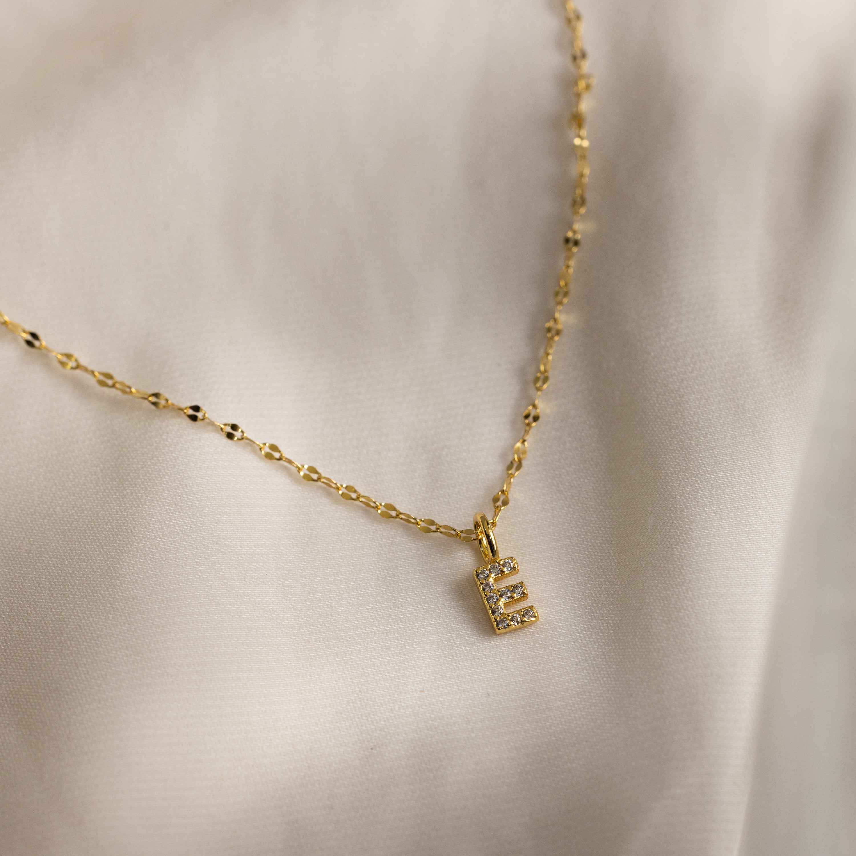 The Dainty Pave Letter Necklace in 18K Gold features an E pendant with sparkling CZ diamonds on a delicate mirror chain, displayed against a cream fabric background.