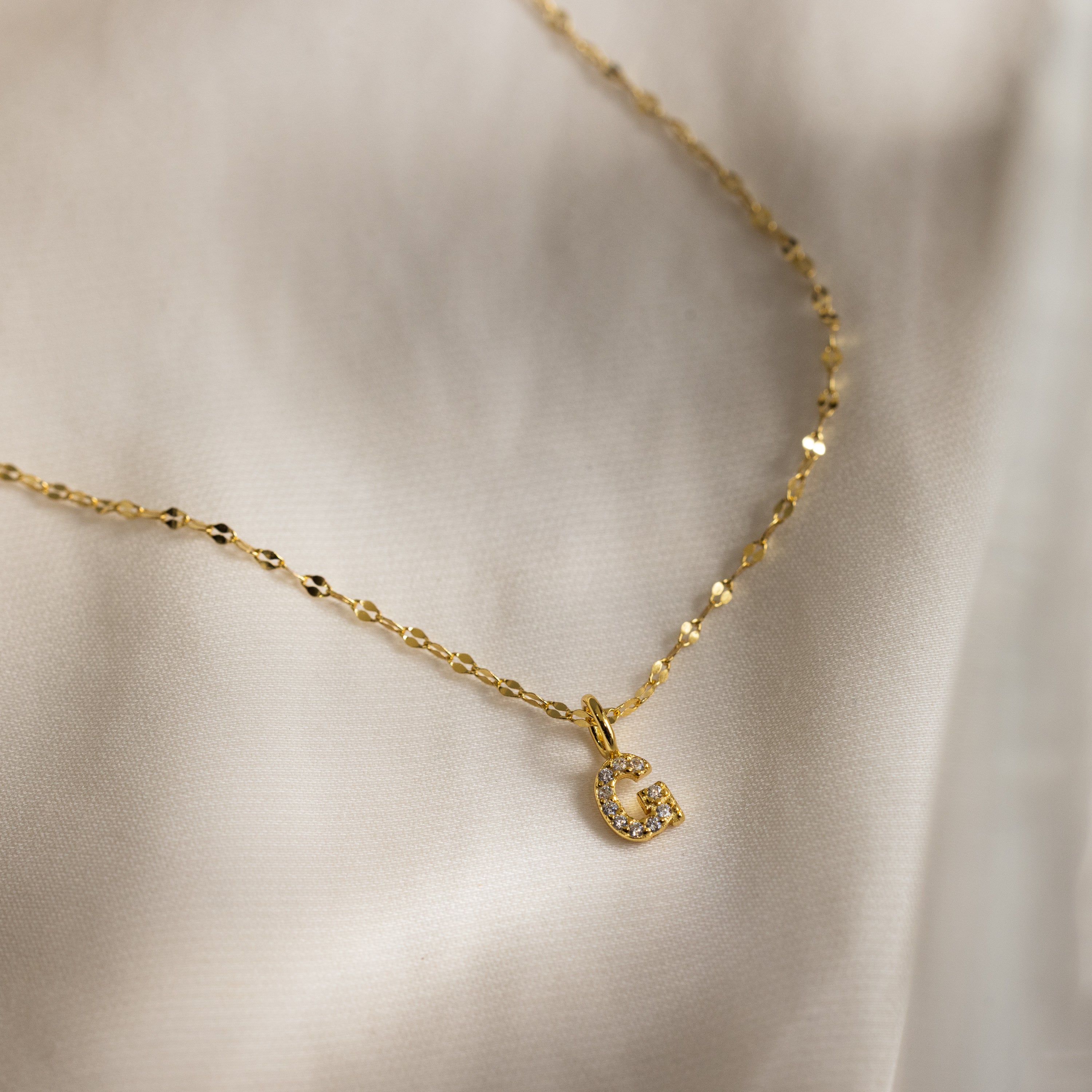 The Dainty Pave Letter Necklace in 18K Gold, featuring a small CZ-studded G pendant, is displayed on a light, silky fabric background.