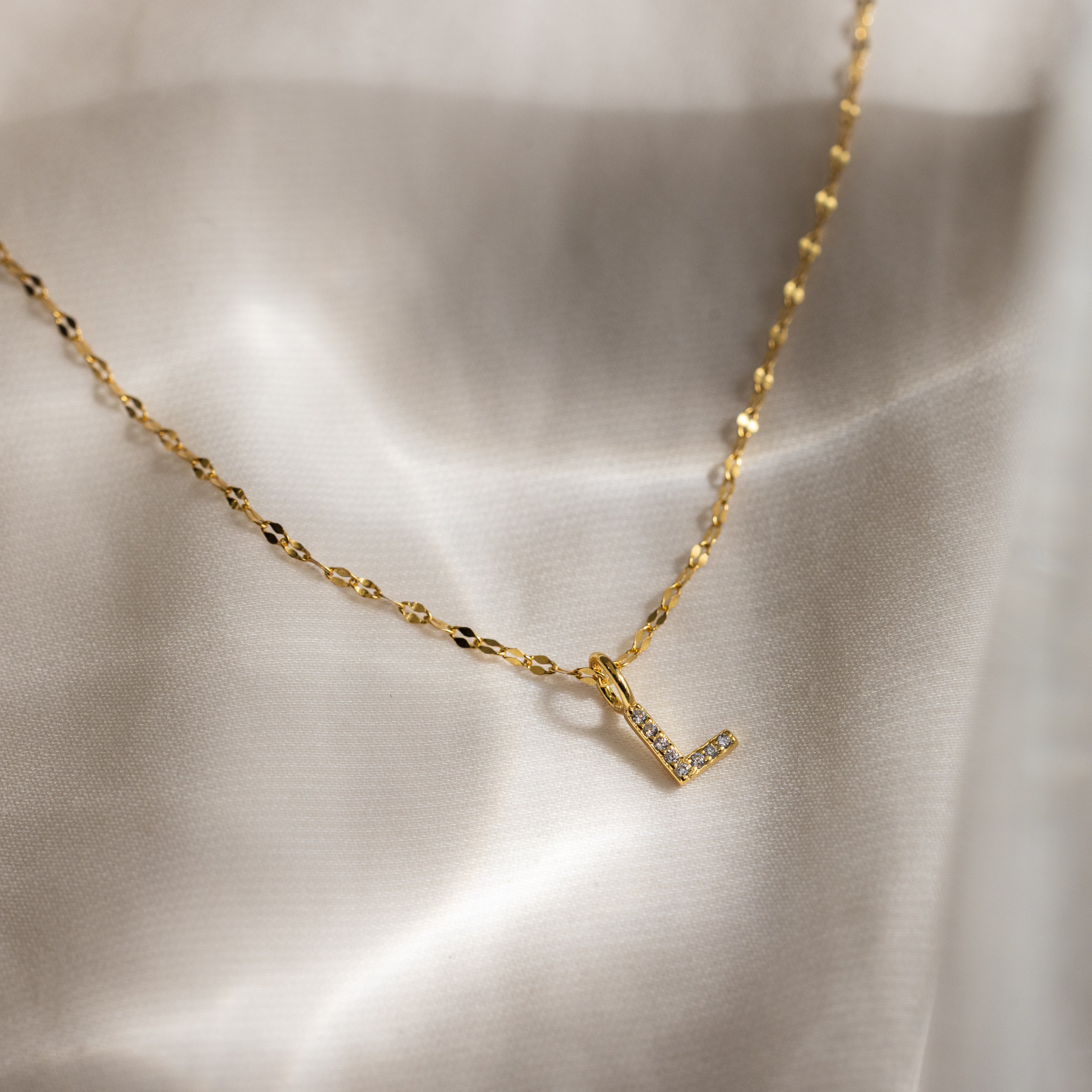 The Dainty Pave Letter Necklace in 18K Gold features a gold mirror chain and a pavé letter L pendant with CZ diamonds, elegantly displayed on silky cream-colored fabric.