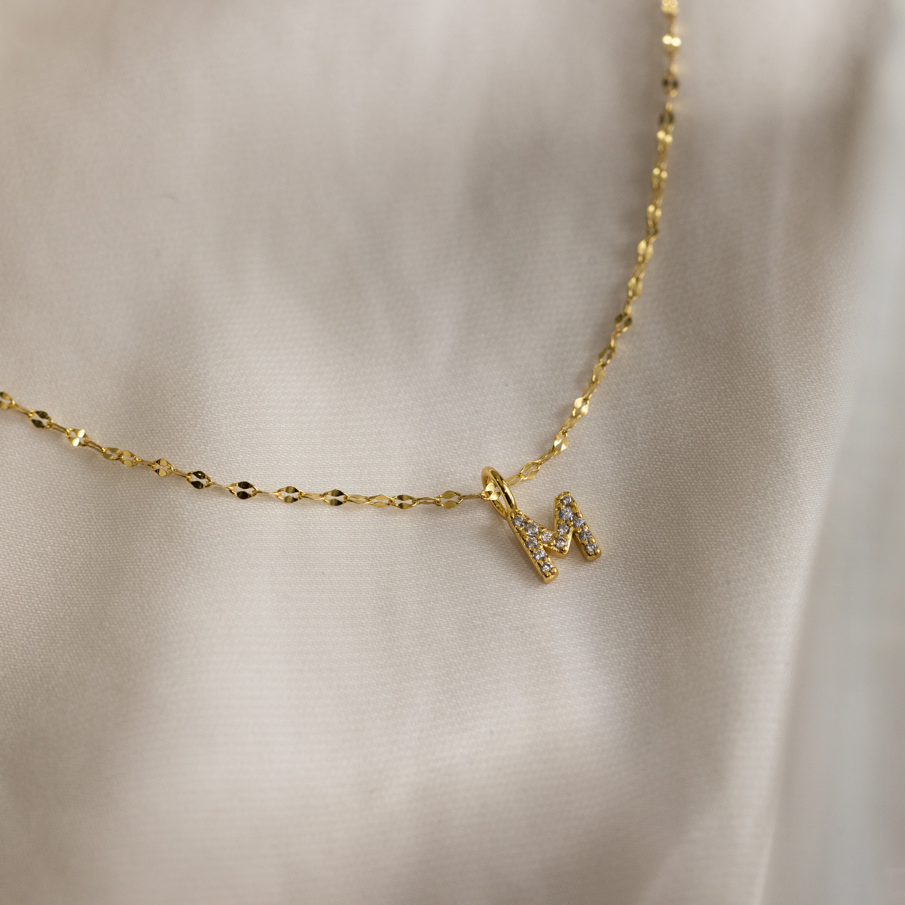 Dainty Pave Letter Necklace in 18K Gold featuring an M pendant encrusted with sparkling CZ diamonds, shown on soft beige fabric.