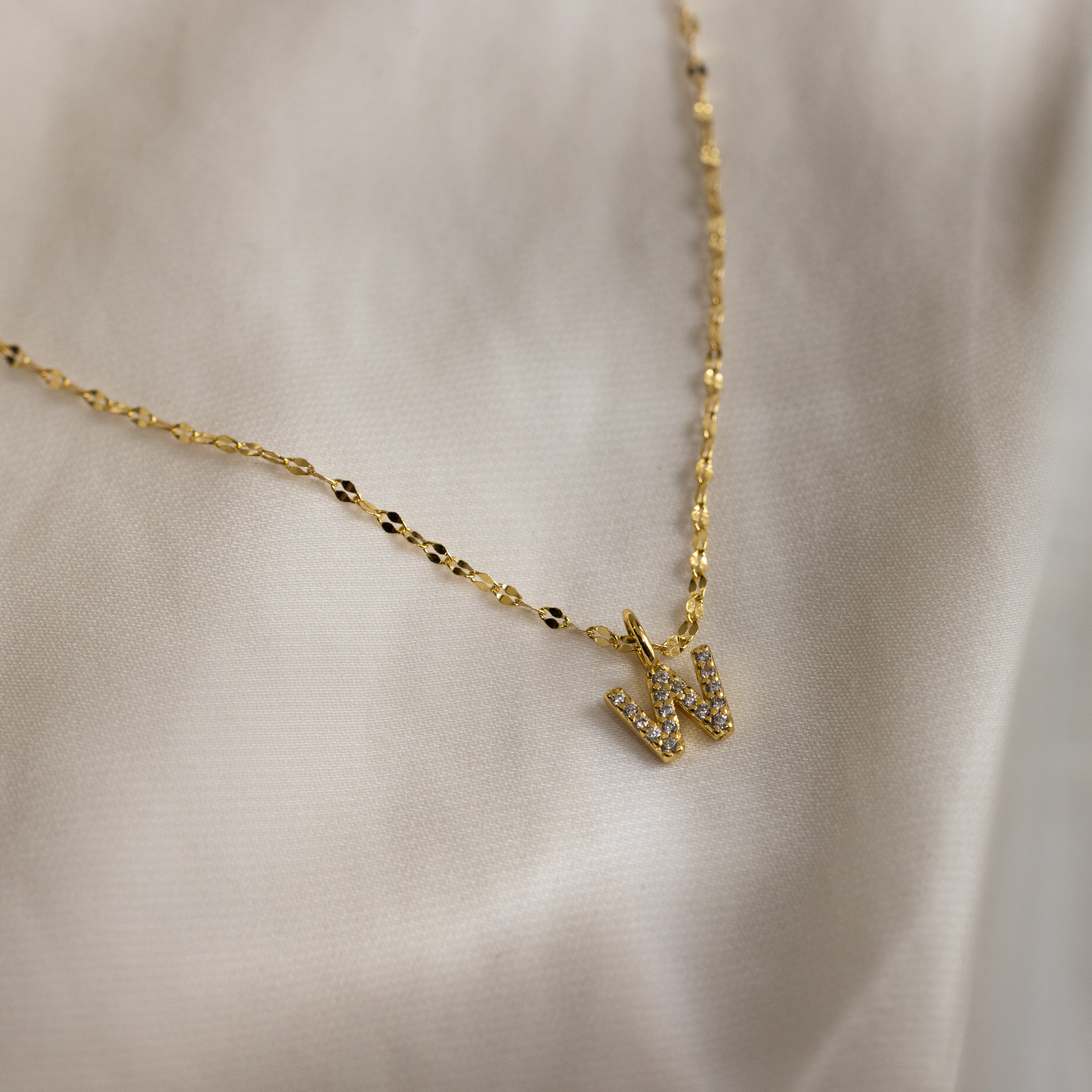 The Dainty Pave Letter Necklace in 18K Gold features a sparkling pavé W pendant with CZ diamonds, shown on a soft, light-colored fabric background.