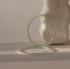 Set of two thin jade bangles in soft translucent green tones, with the darker of the two resting up-right against a stone prop while the lighter bracelet lays flat.