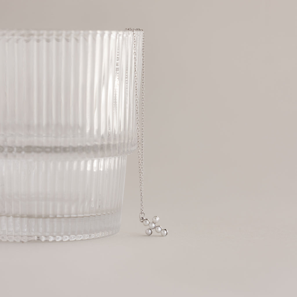The Pearl Cross Necklace in Silver, featuring a delicate pendant, is displayed on a clear ribbed glass against a neutral background.