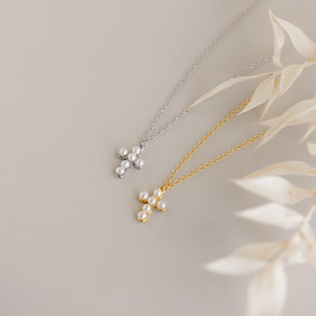 Two Pearl Cross Necklaces—one in silver, one in gold—adorned with luminous pearls are displayed on a light surface with dried leaf accents.