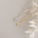 Two Pearl Cross Necklaces—one in silver, one in gold—adorned with luminous pearls are displayed on a light surface with dried leaf accents.