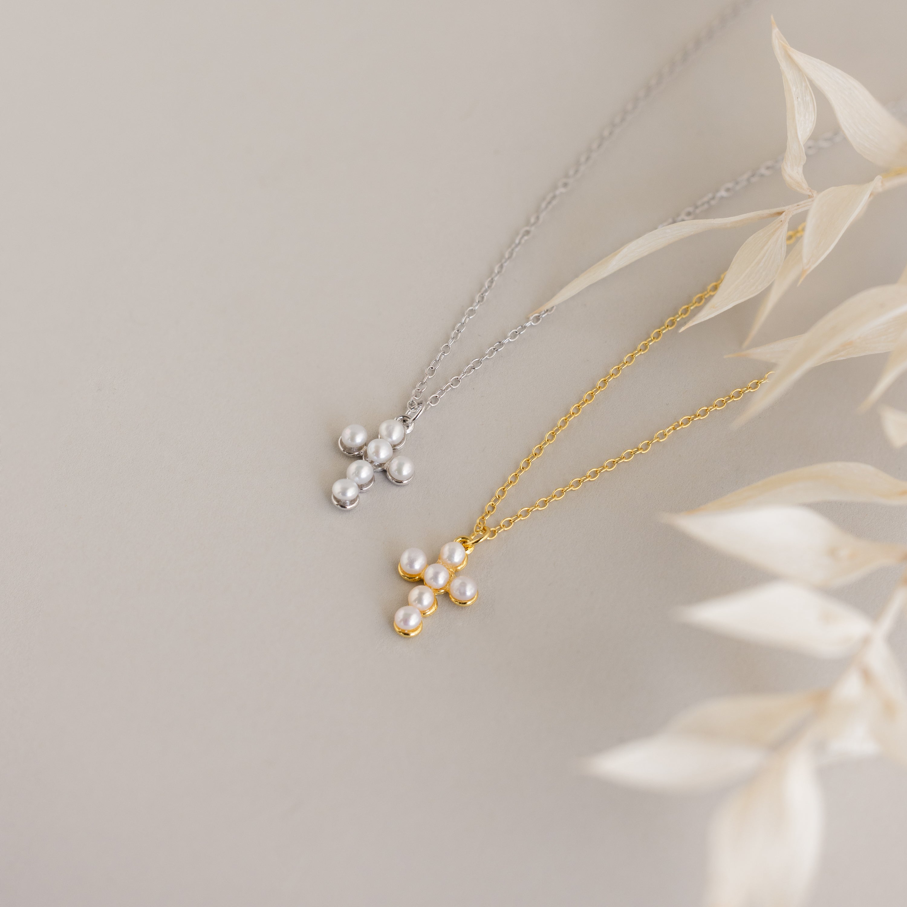 Two Pearl Cross Necklaces—one in silver, one in gold—adorned with luminous pearls are displayed on a light surface with dried leaf accents.