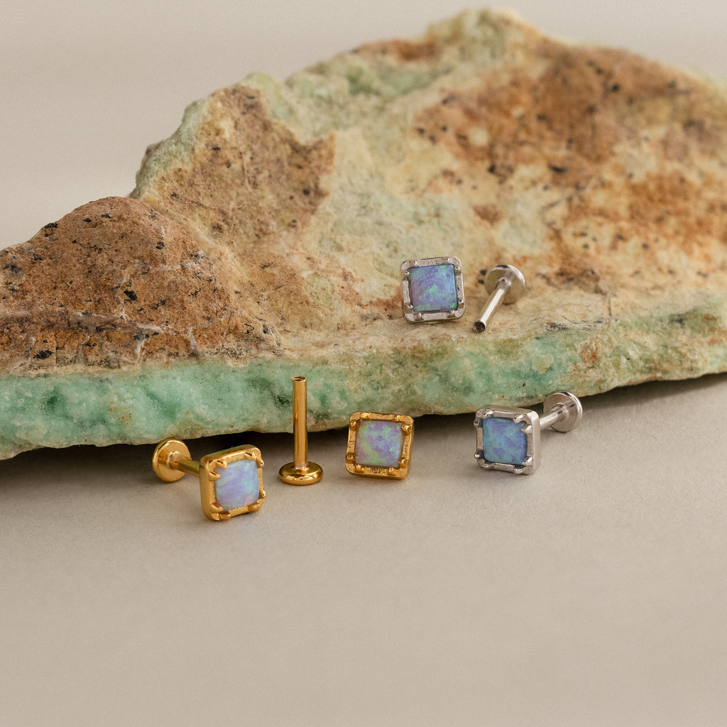 Blue Opal Prism Studs: Three pairs of square blue opal earrings in gold and silver, featuring iridescent stones, displayed on a textured rock.