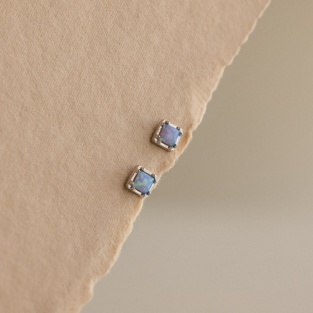 Blue Opal Prism Studs in Silver are displayed on a textured beige surface, reflecting the captivating charm of iridescent jewelry with every glance.