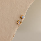 Two Blue Opal Prism Studs in 18K Gold rest on a beige textured surface, adding shimmering elegance and a pop of color to your jewelry collection.