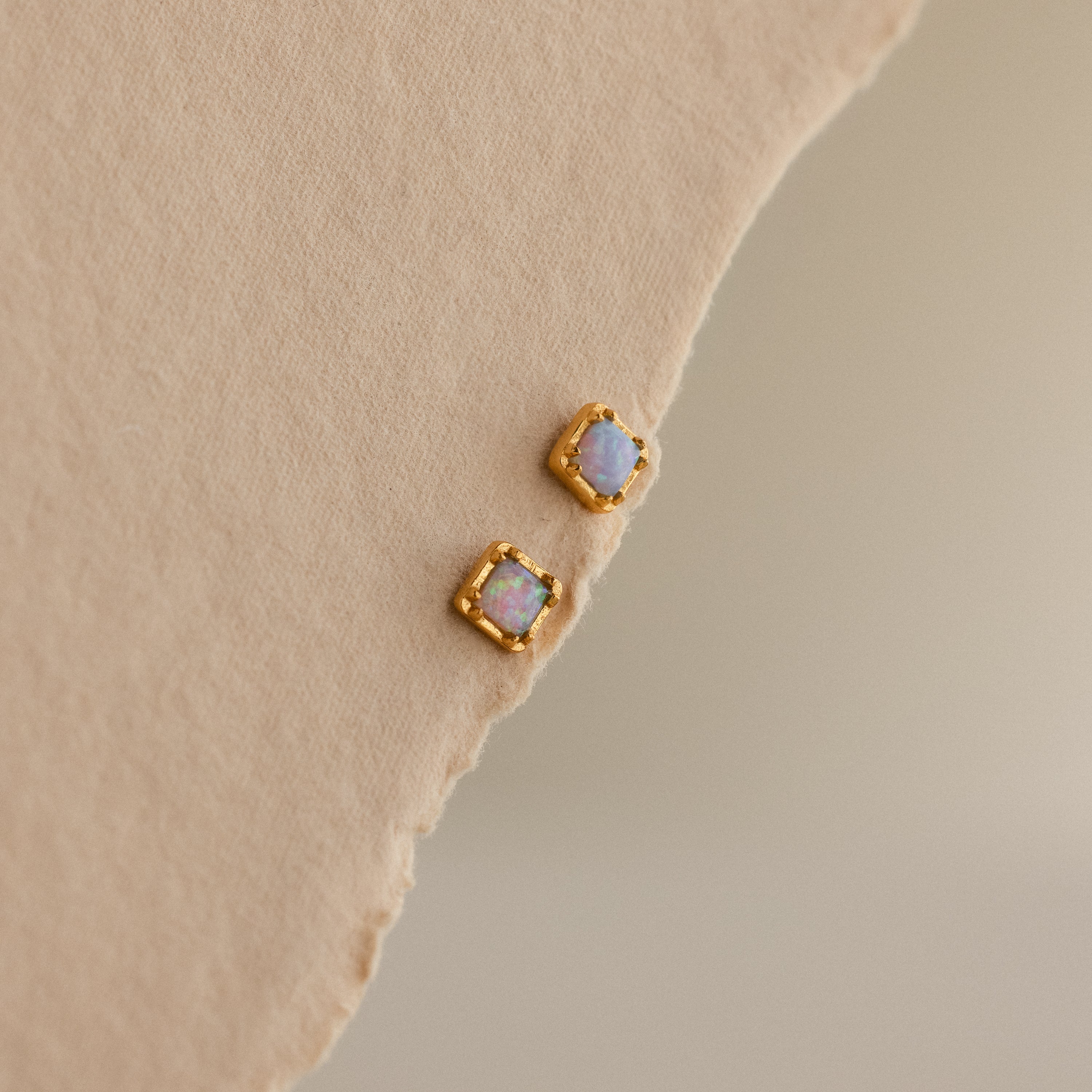 Two Blue Opal Prism Studs in 18K Gold rest on a beige textured surface, adding shimmering elegance and a pop of color to your jewelry collection.