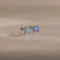Blue Opal Prism Studs in Silver feature square blue opal gemstones that shimmer on a smooth, light beige surface—an elegant, iridescent touch for everyday wear.