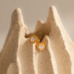 Sacred Heart Studs in 18K Gold with clear gemstones and a subtle sunburst texture are displayed on a textured, light-colored stone surface.