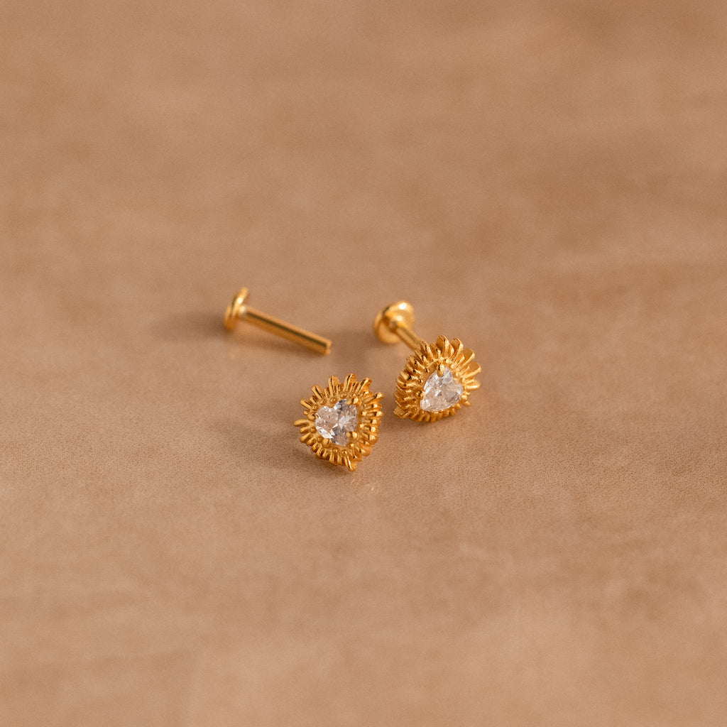 Sacred Heart Studs in 18K Gold, featuring a central clear gem and sunburst texture, displayed on a beige surface.