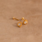 Sacred Heart Studs in 18K Gold, featuring a central clear gem and sunburst texture, displayed on a beige surface.