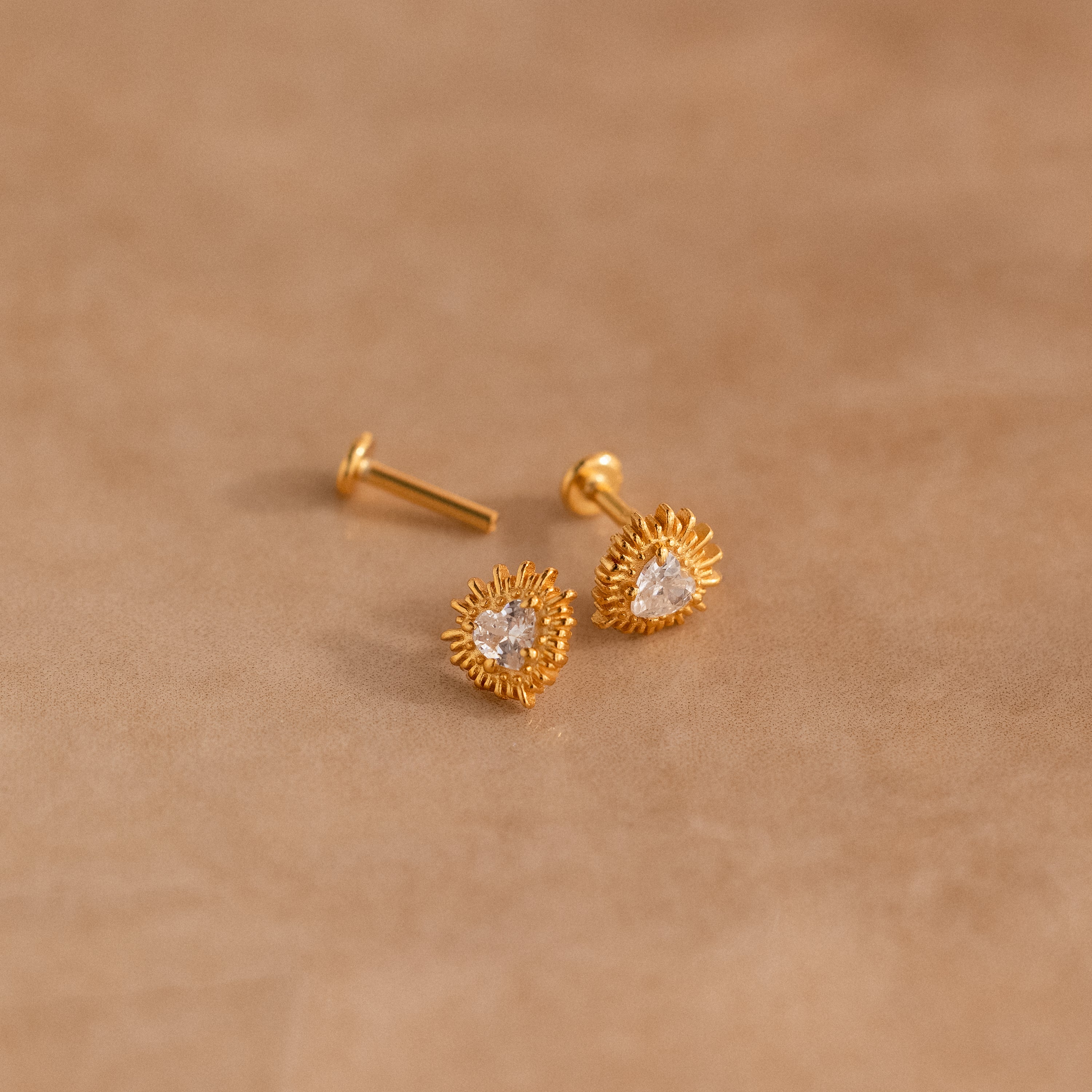 Sacred Heart Studs in 18K Gold, featuring a central clear gem and sunburst texture, displayed on a beige surface.