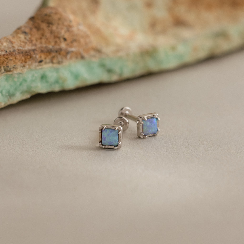 Blue Opal Prism Studs in Silver are displayed on a neutral surface, highlighting their iridescent charm, with a matching stone featured in the background.