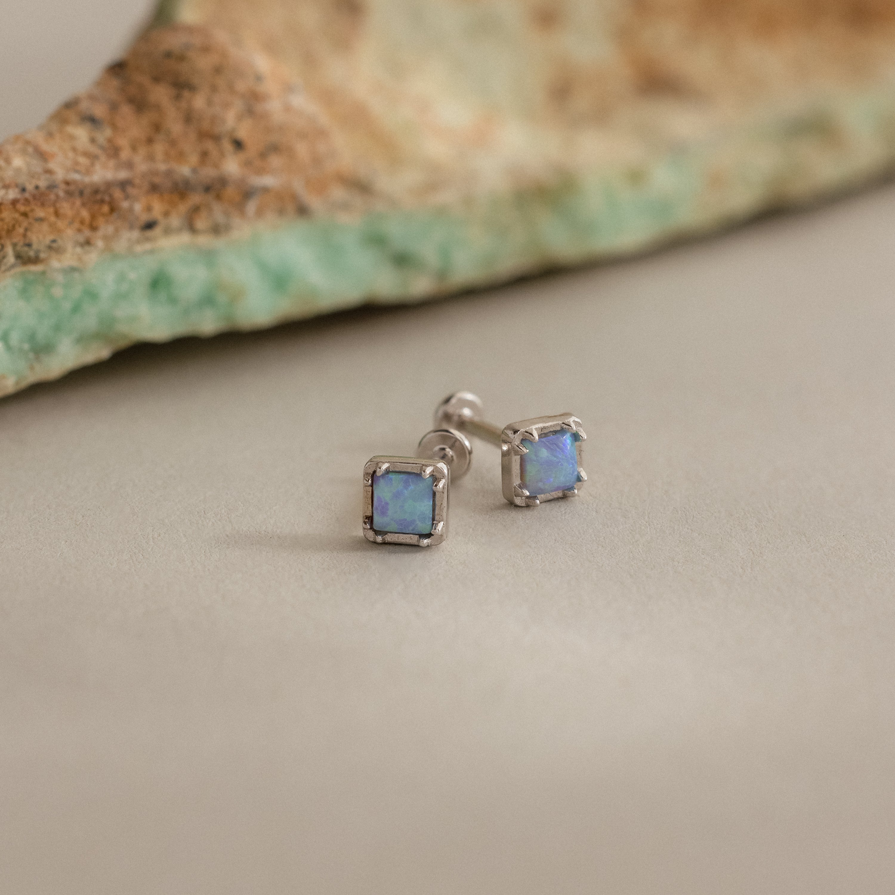 Blue Opal Prism Studs in Silver are displayed on a neutral surface, highlighting their iridescent charm, with a matching stone featured in the background.