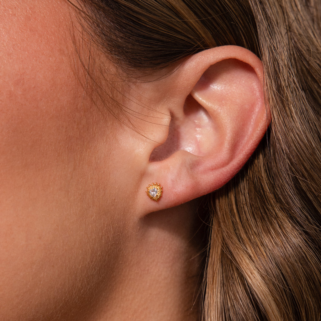 Close-up of a womans ear wearing Sacred Heart Studs in 18K Gold, featuring a sparkling clear gemstone and delicate sunburst texture.