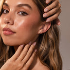 A woman with wavy hair touches her face, showing glossy makeup, nude nails, and Sacred Heart Studs in 18K Gold featuring a stunning sunburst texture.