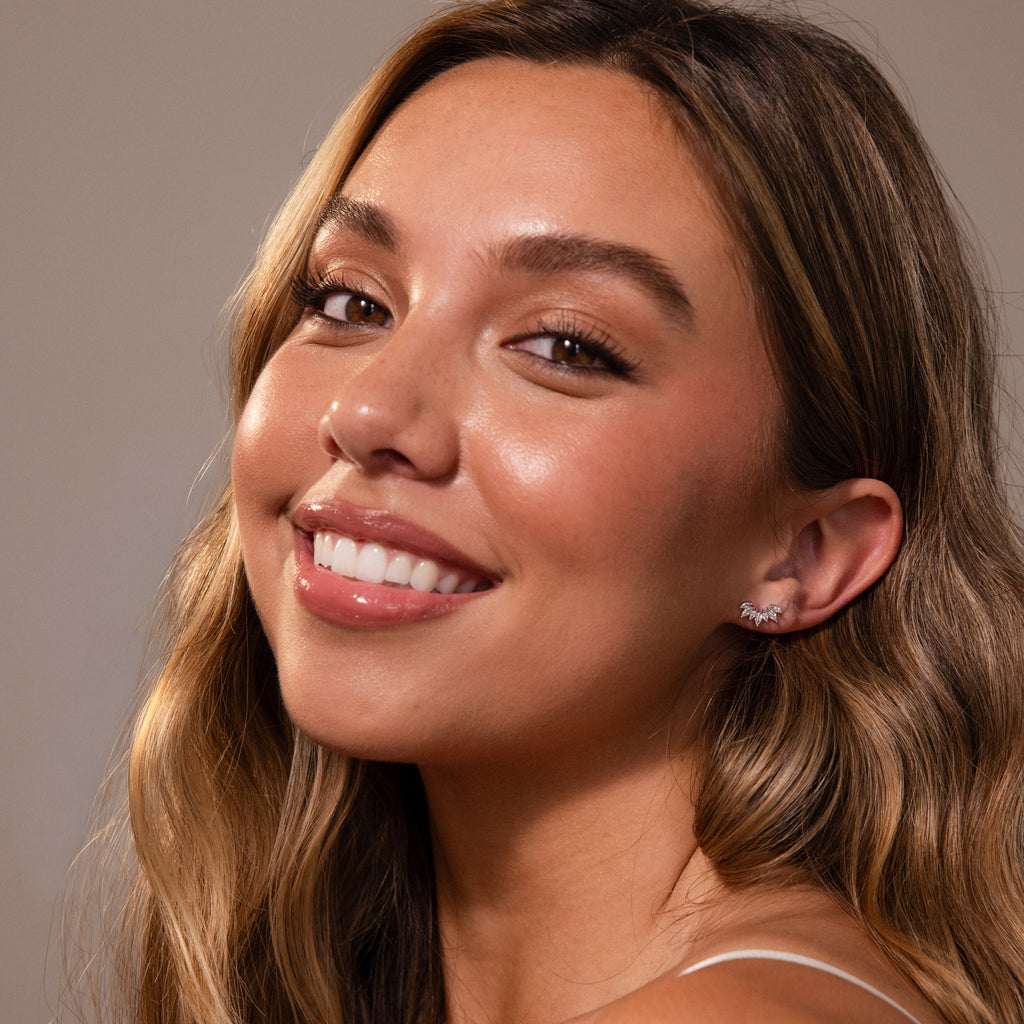A woman with long wavy hair and glowing skin smiles at the camera, elegantly showcasing Marquise Diamond Arc Climbers in Silver against a neutral backdrop.