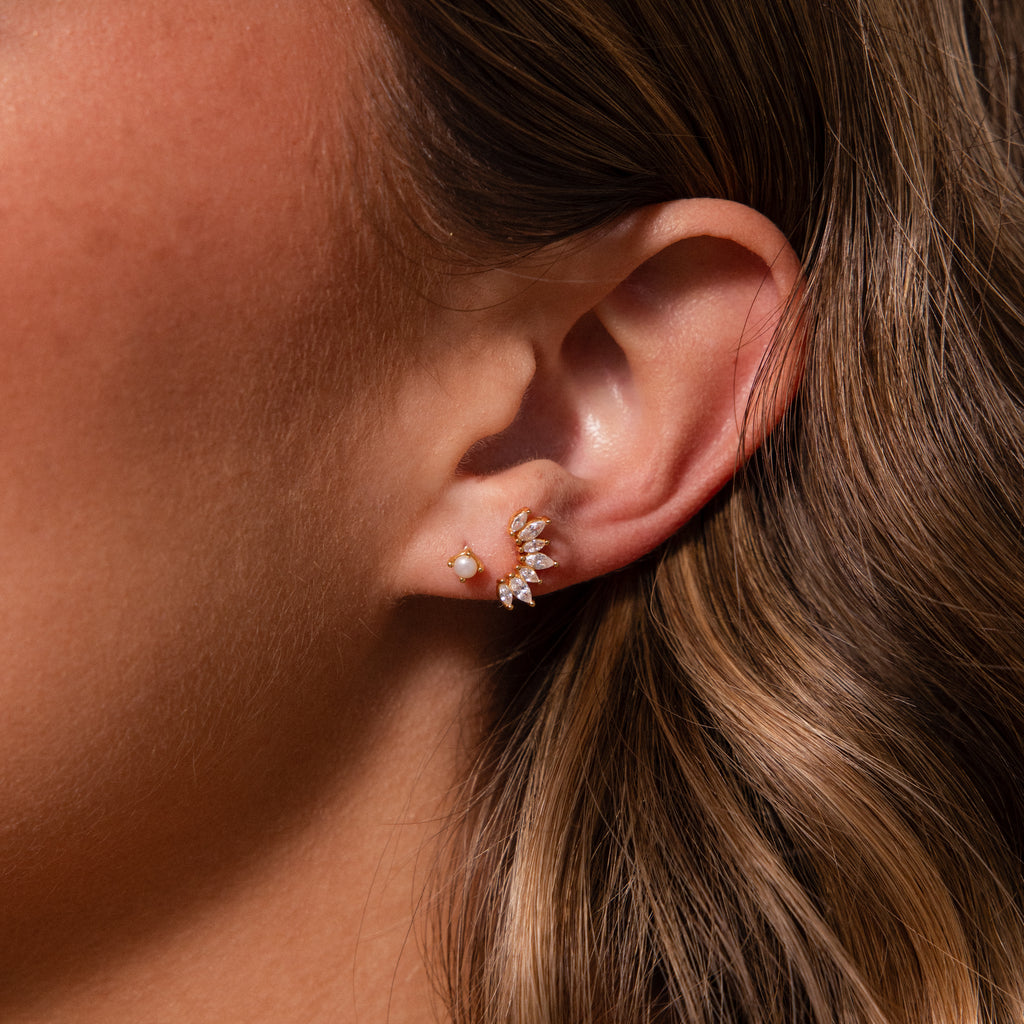 Close-up of a woman’s ear with two earrings: a small pearl stud and Marquise Diamond Arc Climbers in 18K Gold, adorned with sparkling marquise stones.