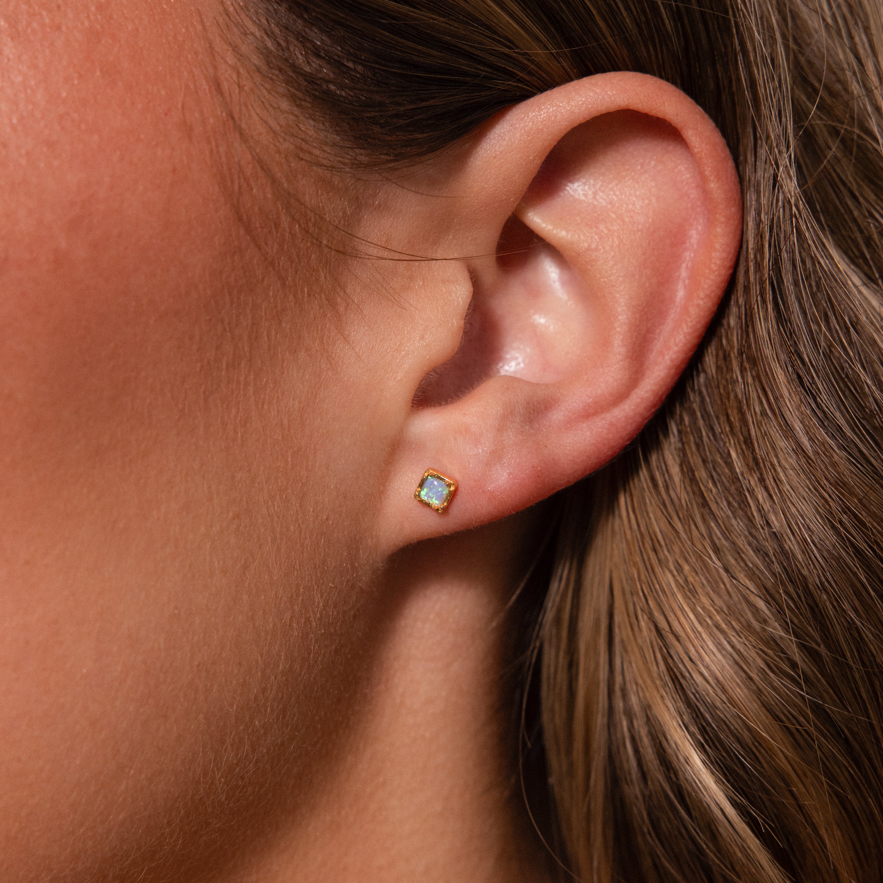 Close-up of a woman’s ear wearing Blue Opal Prism Studs in 18K Gold—small, iridescent square blue opal earrings set in delicate gold.