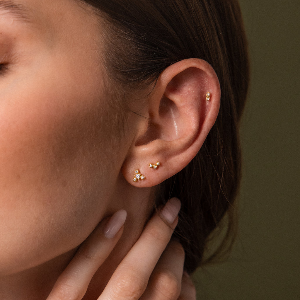 Close-up of a woman's ear wearing small gold stud earrings and a gold helix piercing against a muted olive background.