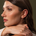 A close-up side profile of a woman with multiple small gold ear piercings and earrings, including a small hoop in her lobe and studs on her ear cartilage, against a dark olive background.