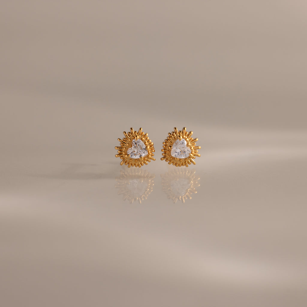Sacred Heart Studs in 18K Gold feature a radiant sunburst texture with clear center stones, shown on a soft neutral background.