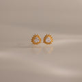 Sacred Heart Studs in 18K Gold feature a radiant sunburst texture with clear center stones, shown on a soft neutral background.
