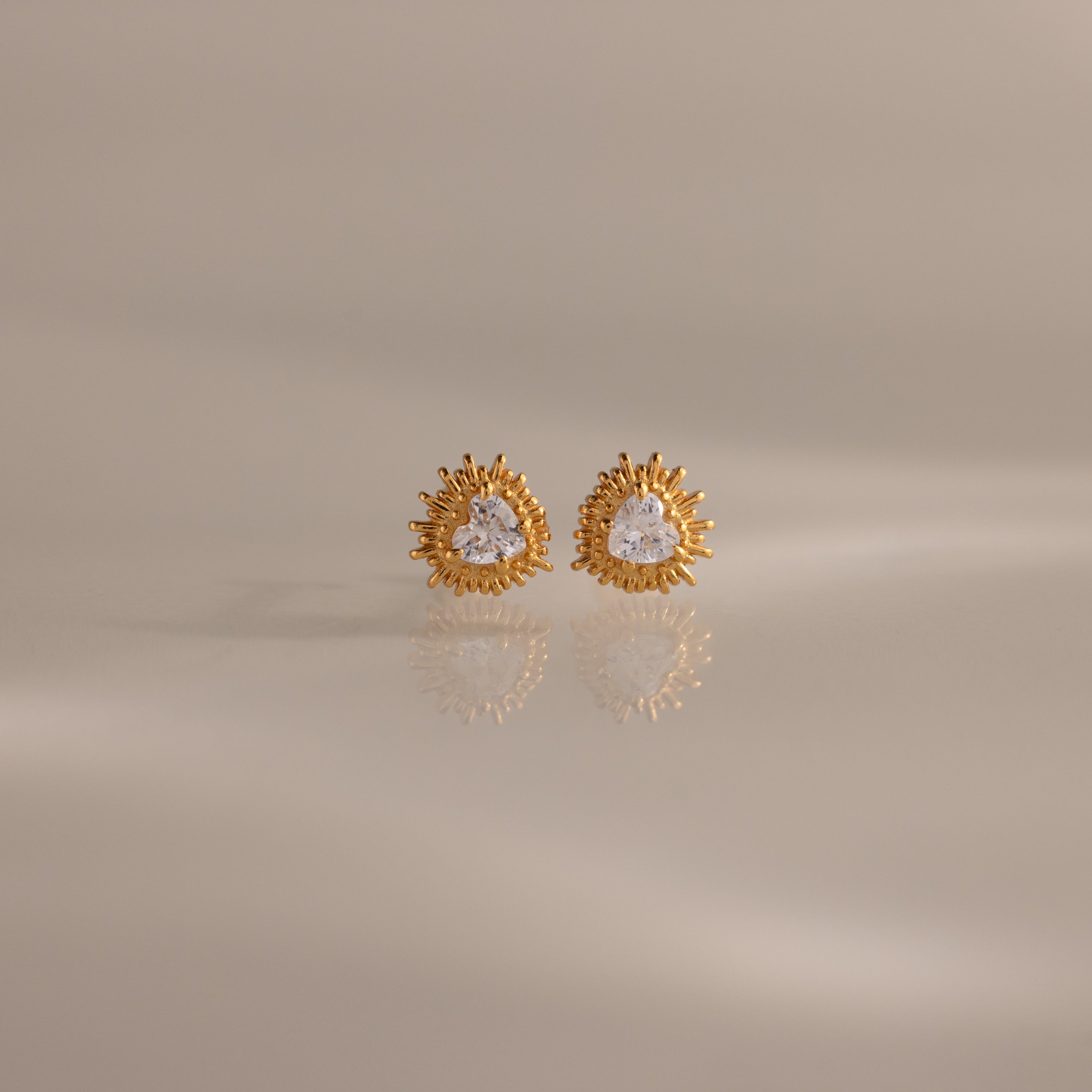 Sacred Heart Studs in 18K Gold feature a radiant sunburst texture with clear center stones, shown on a soft neutral background.