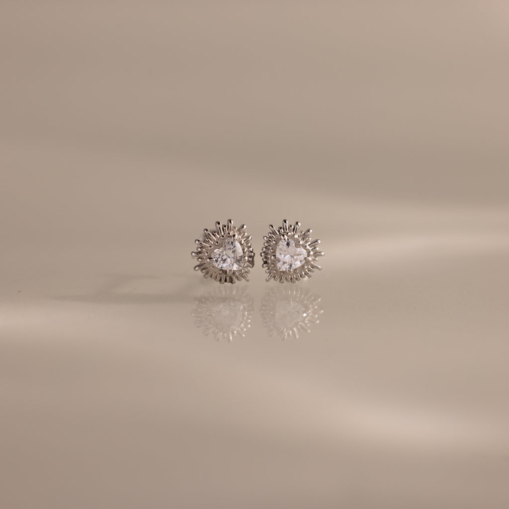 The Sacred Heart Studs in Silver feature round sparkling stones and a sunburst texture, shown on a smooth beige surface.