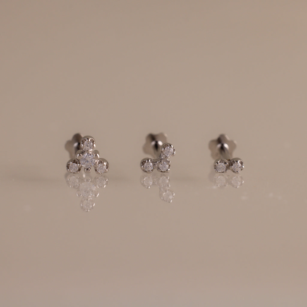 Three pairs of silver stud earrings with small clear gemstones, arranged on a reflective beige surface against a solid beige background.