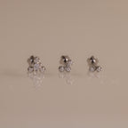 Three pairs of silver stud earrings with small clear gemstones, arranged on a reflective beige surface against a solid beige background.