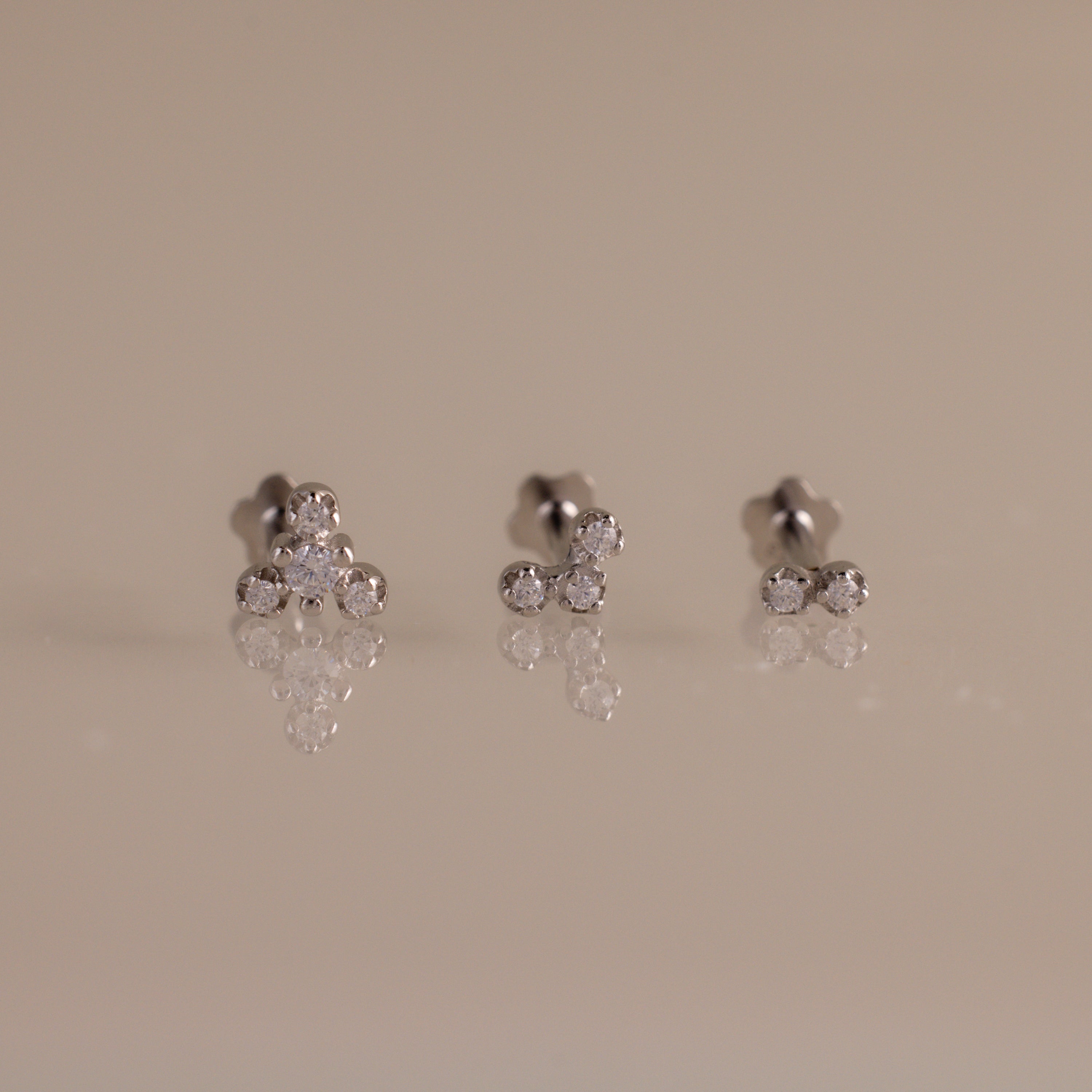 Three pairs of silver stud earrings with small clear gemstones, arranged on a reflective beige surface against a solid beige background.