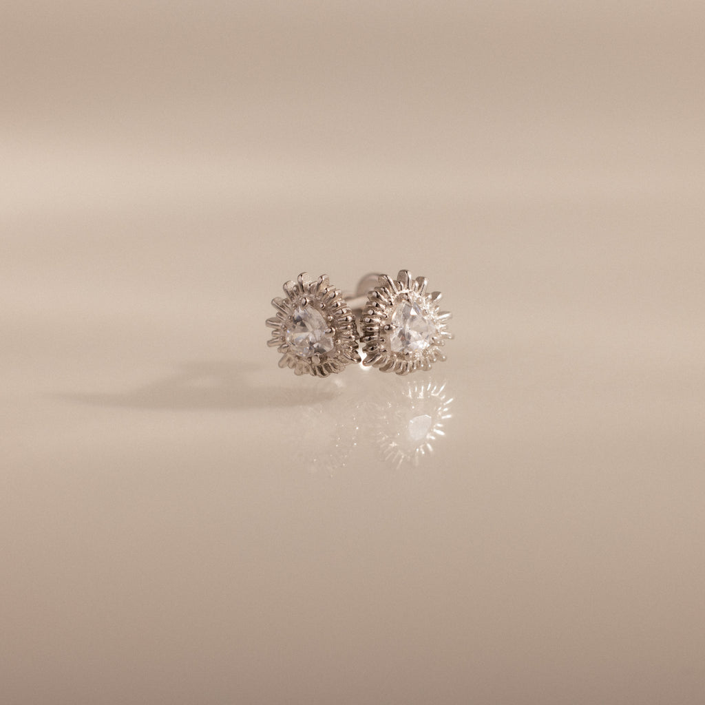 Sacred Heart Studs in Silver featuring round clear gemstones and a sunburst texture, displayed on a soft beige background.