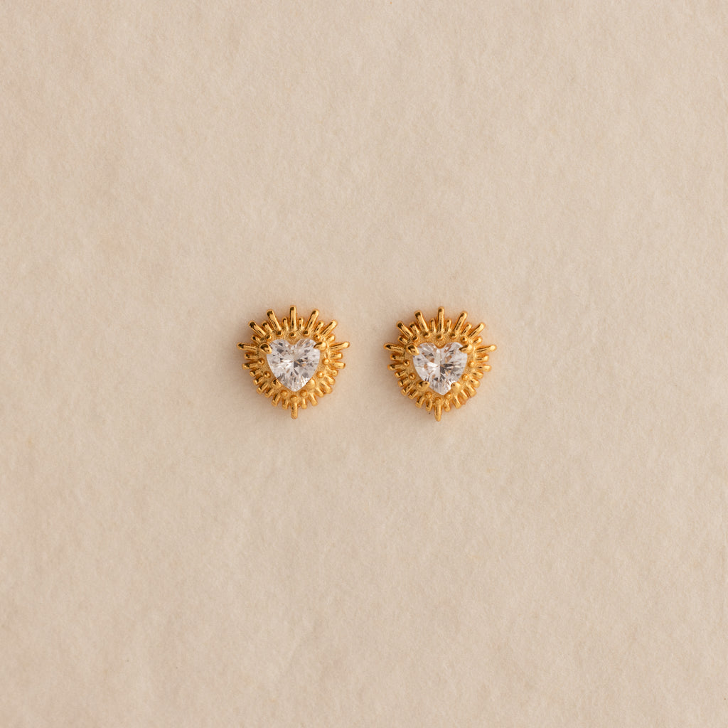 Sacred Heart Studs in 18K Gold, featuring a heart-shaped center stone and radiant sunburst texture, displayed on a beige background.