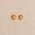 Sacred Heart Studs in 18K Gold, featuring a heart-shaped center stone and radiant sunburst texture, displayed on a beige background.