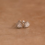 The Sacred Heart Studs in Silver, featuring teardrop-shaped clear stones and a sunburst texture, add a radiant touch as they rest on a soft brown surface.