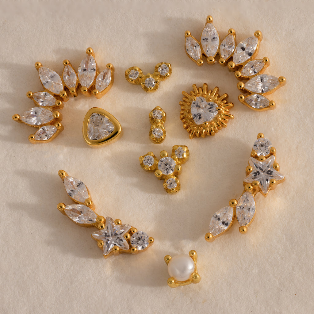 Assorted gold crystal flat back earrings featuring marquise, star, heart, pearl, and cluster designs arranged on a neutral background.
