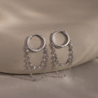The Duo Mirror Chain Huggies in Sterling Silver feature two delicate chain strands and are shown on a smooth beige surface—ideal for those who love modern jewelry.